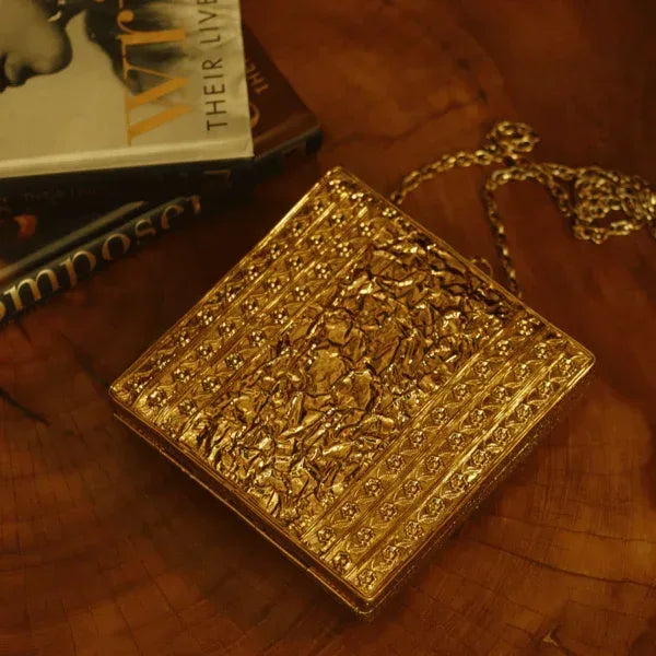 A brass clutch purse with intricate embellishments, featuring a sleek design and decorative details, by The Tan Clan.